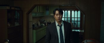 Movie still from “Decision to Leave” (2022), directed by Park Chan-wook – A man wearing a suit and tie standing in a kitchen; Medium shot, Low angle