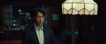 Movie still from “Decision to Leave” (2022), directed by Park Chan-wook – A man standing in front of a lamp in a room; Medium shot, Over the shoulder angle