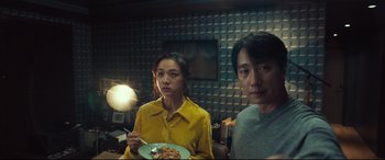 Movie still from “Decision to Leave” (2022), directed by Park Chan-wook – A woman holding a plate of food next to a man; Medium shot, Over the shoulder angle