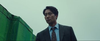 Movie still from “Decision to Leave” (2022), directed by Park Chan-wook – A man in a suit and tie standing in front of a green building; Close Up shot, Low angle