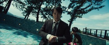 Movie still from “Decision to Leave” (2022), directed by Park Chan-wook – A man wearing a suit and tie standing next to a tree; Medium shot, Low angle