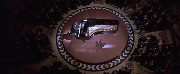 Movie still from “Gattaca” (1997), directed by Andrew Niccol – A person playing a grand piano in a circle of people; Extreme Wide shot, Overhead angle