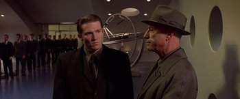 Movie still from “Gattaca” (1997), directed by Andrew Niccol – A man in a suit standing next to another man in a hat; Medium shot, Over the shoulder angle