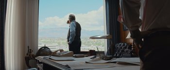 Movie still from “Gold” (2016), directed by Stephen Gaghan – An older man standing in front of a large window; Wide shot, Over the shoulder angle