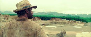 Movie still from “Gold” (2016), directed by Stephen Gaghan – A man wearing a hat looking out over a dirt field; Medium shot, Over the shoulder angle