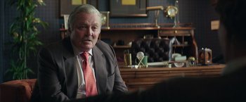 Movie still from “Gold” (2016), directed by Stephen Gaghan – An older man sitting in a chair in front of a desk; Close Up shot, Over the shoulder angle