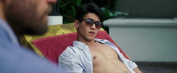 Movie still from “Gold” (2016), directed by Stephen Gaghan – A shirtless man wearing sunglasses sitting on top of a couch; Medium shot, Over the shoulder angle