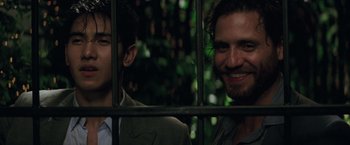 Movie still from “Gold” (2016), directed by Stephen Gaghan – Two men are smiling behind a metal fence; Close Up shot, Over the shoulder angle