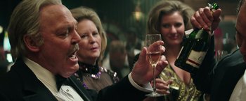 Movie still from “Gold” (2016), directed by Stephen Gaghan – A man holding a glass of wine in front of two other people; Close Up shot, Low angle