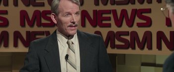 Movie still from “Gold” (2016), directed by Stephen Gaghan – A man in a suit and tie standing in front of a sign; Close Up shot, Over the shoulder angle