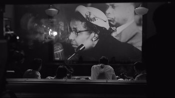 Movie still from “Good Night, and Good Luck.” (2005), directed by George Clooney – People are sitting in front of a large picture of a man; Medium shot, Over the shoulder angle