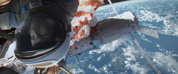 Movie still from “Gravity” (2013), directed by Alfonso Cuarón – A person is holding a paper in his hand while standing on the side of a space station; Extreme Wide shot, Overhead angle