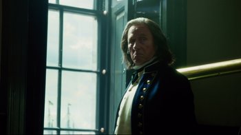 Movie still from “In the Heart of the Sea” (2015), directed by Ron Howard – A man in a blue jacket standing in front of a window; Medium shot, Over the shoulder angle