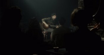Movie still from “Inside Llewyn Davis” (2013), directed by Ethan Coen – A man sitting in a chair playing a guitar; Wide shot, Over the shoulder angle