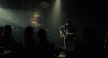 Movie still from “Inside Llewyn Davis” (2013), directed by Ethan Coen – A man playing an acoustic guitar in a dark room; Wide shot, Over the shoulder angle