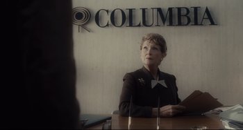 Movie still from “Inside Llewyn Davis” (2013), directed by Ethan Coen – An older woman sitting at a table in front of a columbia sign; Medium shot, Over the shoulder angle
