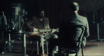 Movie still from “Inside Llewyn Davis” (2013), directed by Ethan Coen – A man playing an acoustic guitar while another man sits in a chair; Medium shot, Over the shoulder angle