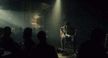 Movie still from “Inside Llewyn Davis” (2013), directed by Ethan Coen – A man sitting on a chair playing a guitar; Wide shot, Over the shoulder angle