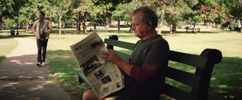 Movie still from “Irrational Man” (2015), directed by Woody Allen – An older man sitting on a park bench reading a newspaper; Medium shot, Over the shoulder angle