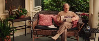 Movie still from “Irrational Man” (2015), directed by Woody Allen – An older man sitting on a wicker couch reading; Medium shot, Over the shoulder angle