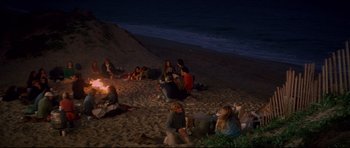 Movie still from “Jaws” (1975), directed by Steven Spielberg – A group of people sitting on top of a sandy beach at night; Wide shot, High angle
