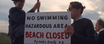 Movie still from “Jaws” (1975), directed by Steven Spielberg – A person holding a sign that reads " no swimming hazardous area beach closed "; Close Up shot, High angle