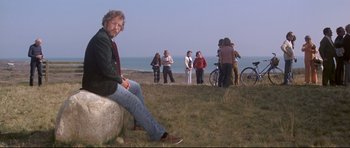 Movie still from “Jaws” (1975), directed by Steven Spielberg – A man sitting on top of a rock in front of a group of people; Wide shot, Low angle