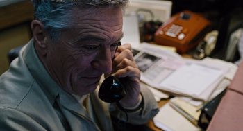 Movie still from “Joy” (2015), directed by David O. Russell – An older man talking on a phone while sitting at a desk; Close Up shot, High angle