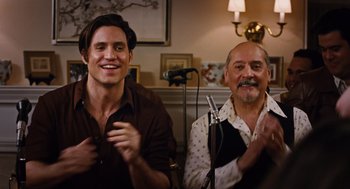 Movie still from “Joy” (2015), directed by David O. Russell – Two men sitting in a living room with microphones; Medium shot, Over the shoulder angle