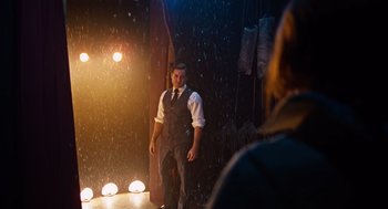 Movie still from “Joy” (2015), directed by David O. Russell – A man standing in front of a stage with lights in the background; Wide shot, Over the shoulder angle