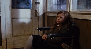 Movie still from “Joy” (2015), directed by David O. Russell – A young girl sitting in a chair in a room; Medium shot, Over the shoulder angle