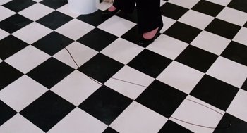 Movie still from “Joy” (2015), directed by David O. Russell – A woman standing on a checkerboard tile floor; Medium shot, Overhead angle