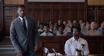 Movie still from “Just Mercy” (2019), directed by Destin Daniel Cretton – A man in a suit and tie in front of a crowd of people in a court room; Medium shot, Over the shoulder angle