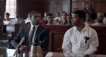 Movie still from “Just Mercy” (2019), directed by Destin Daniel Cretton – Two men in suits sit in front of a group of people in a court room; Medium shot, Over the shoulder angle