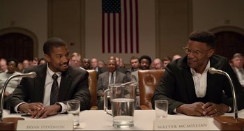Movie still from “Just Mercy” (2019), directed by Destin Daniel Cretton – Two men sitting at a table in front of a large american flag; Medium shot, Over the shoulder angle