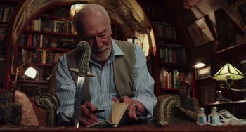 Movie still from “Knives Out” (2019), directed by Rian Johnson – An older man writing in a book while holding a sword; Medium shot, Low angle