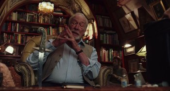 Movie still from “Knives Out” (2019), directed by Rian Johnson – An older man sitting at a table in front of a bookshelf; Medium shot, Over the shoulder angle