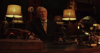 Movie still from “Knives Out” (2019), directed by Rian Johnson – An older man sitting at a desk in front of a lamp; Medium shot, Low angle