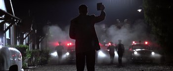 Movie still from “L.A. Confidential” (1997), directed by Curtis Hanson – A man in a black suit holding up a sign in front of a line of police cars at night; Medium shot, Low angle