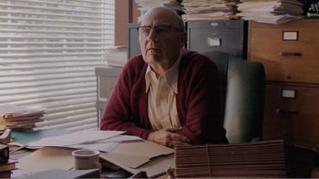 Movie still from “Don't Worry, He Won't Get Far on Foot” (2018), directed by Gus Van Sant – An older man sitting at a desk in front of a stack of papers; Medium shot, Over the shoulder angle