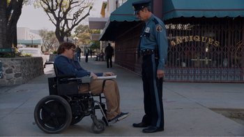 Movie still from “Don't Worry, He Won't Get Far on Foot” (2018), directed by Gus Van Sant – A man in a wheel chair talking to a police officer on the sidewalk; Wide shot, High angle