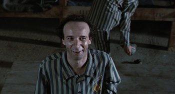 Movie still from “Life Is Beautiful” (1997), directed by Roberto Benigni – A man in striped pajamas smiling for the camera; Medium shot, Over the shoulder angle