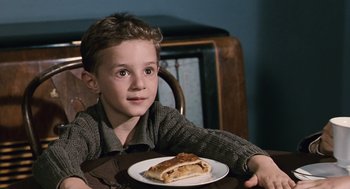 Movie still from “Life Is Beautiful” (1997), directed by Roberto Benigni – A boy sitting at a table with a piece of cake on a plate; Close Up shot, Over the shoulder angle