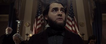 Movie still from “Lincoln” (2012), directed by Steven Spielberg – A man with a beard wearing a suit and bow tie in front of american flags; Close Up shot, Low angle