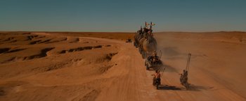 Movie still from “Mad Max: Fury Road” (2015), directed by George Miller – A group of people riding on the back of a train on the side of a dirt road; Extreme Wide shot, High angle