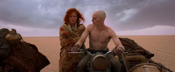 Movie still from “Mad Max: Fury Road” (2015), directed by George Miller – A man riding a motorcycle in the desert with a woman; Medium shot, Low angle