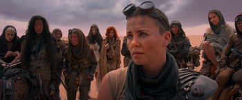 Movie still from “Mad Max: Fury Road” (2015), directed by George Miller – A group of people standing on top of a dirt field; Medium shot, Over the shoulder angle