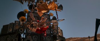 Movie still from “Mad Max: Fury Road” (2015), directed by George Miller – A musical instrument is hanging from the side of a building; Wide shot, Low angle