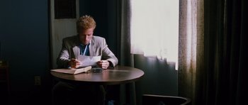 Movie still from “Memento” (2000), directed by Christopher Nolan – A man sitting at a table with papers and a calculator; Medium shot, Over the shoulder angle