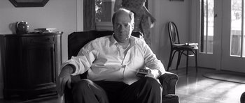 Movie still from “Memento” (2000), directed by Christopher Nolan – A black and white photo of a man sitting in a chair holding a tv remote; Medium shot, Low angle